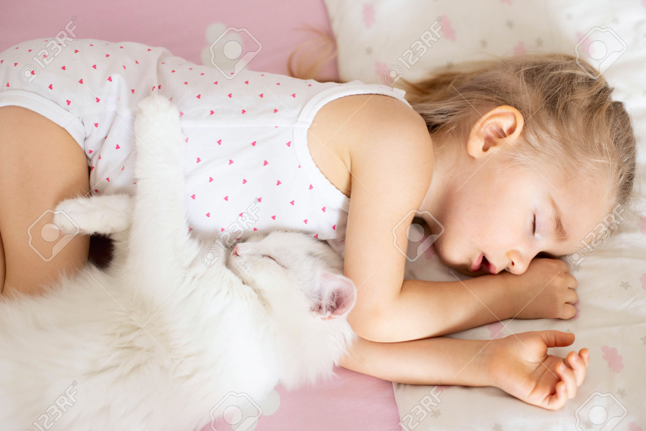 A little girl sleeps in a crib with pink bed linen with clouds, a white cat in the nursery. Quiet hour, top view. - 167408261 A little girl sleeps in a crib with pink bed linen with clouds, a white cat in the nursery. Quiet hour, top view. - 167408261