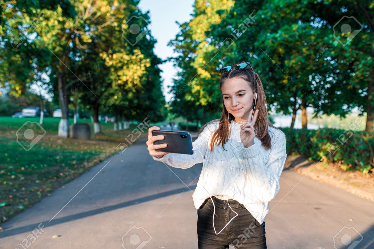 NATU街撮り女子高生　 10代の少女の女子高生、自分で写真を撮る10代のヘッドフォン、電話、楽しんで幸せな笑顔、ビデオの録画、ビデオ通話、秋の夏の街、白いセーター、ジーンズ。背景の木の道の写真素材・画像素材  Image 133744318