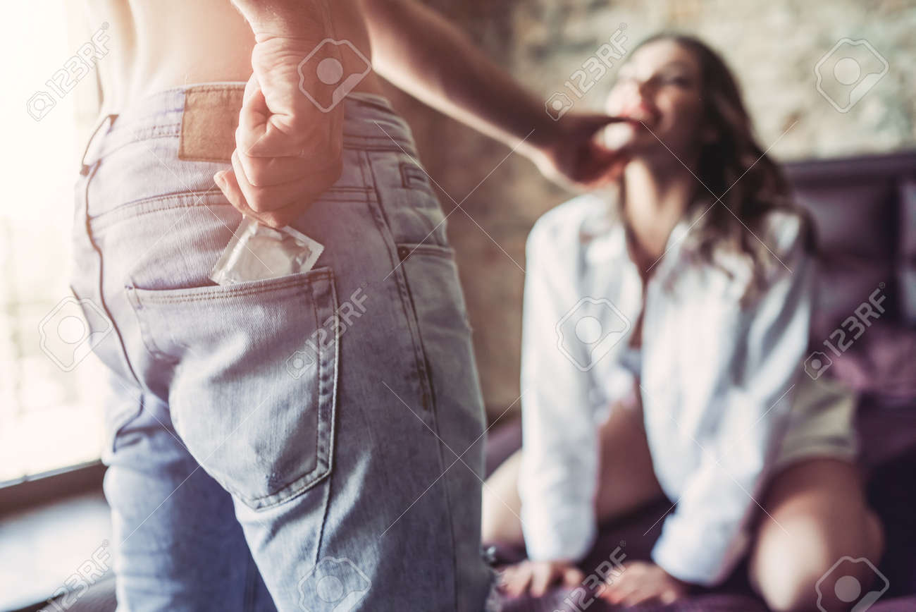 女性はバック グラウンドでのベッドの上に座っている間 ジーンズからコンドームを取って男の画像をトリミングしました 情熱的なカップルの寝室でセックスです の写真素材 画像素材 Image