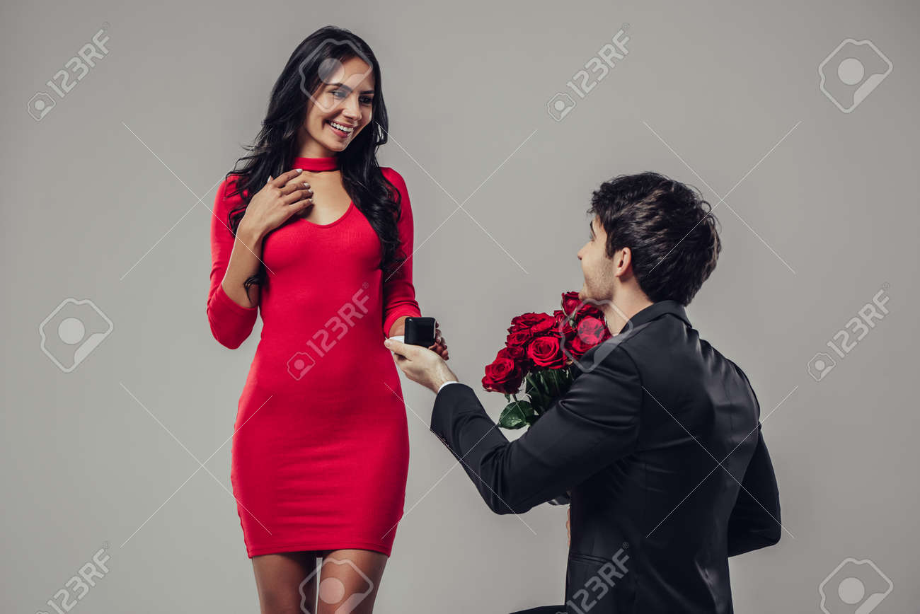 Lindo Par Romantico Isolado Em Fundo Cinza Homem Bonito De Terno Esta Fazendo Proposta Para Sua Jovem Mulher Atraente No Vestido Vermelho Casal Apaixonado Feliz Dia Dos Namorados Fotos Retratos Imagenes Y