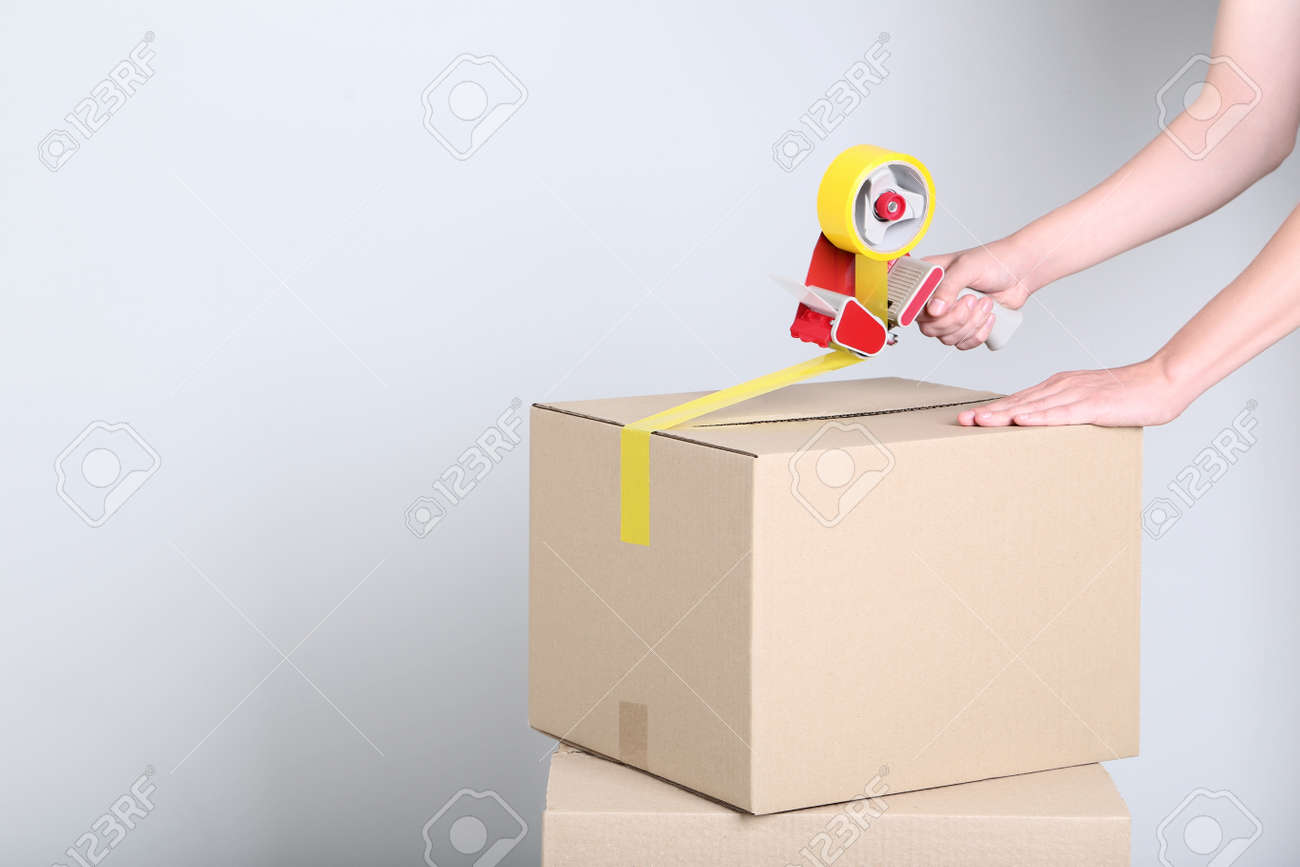 Female's hands packaging cardboard box with dispenser on grey background - 113295005 Female's hands packaging cardboard box with dispenser on grey background - 113295005