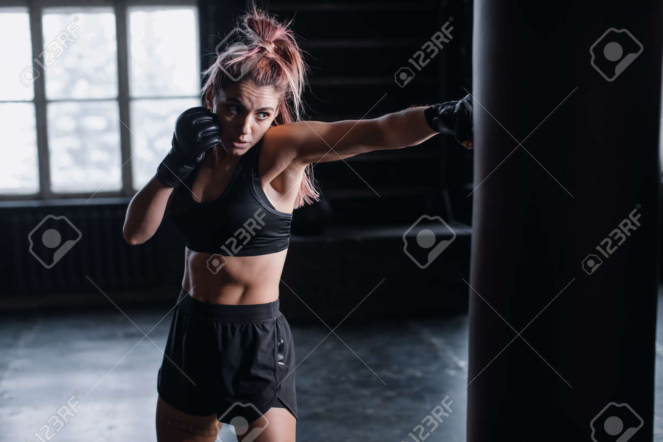 Athletic fit female boxer exercising punches with boxing bag in gym during kickboxing and self defense intensive workout - 166782006 Athletic fit female boxer exercising punches with boxing bag in gym during kickboxing and self defense intensive workout - 166782006