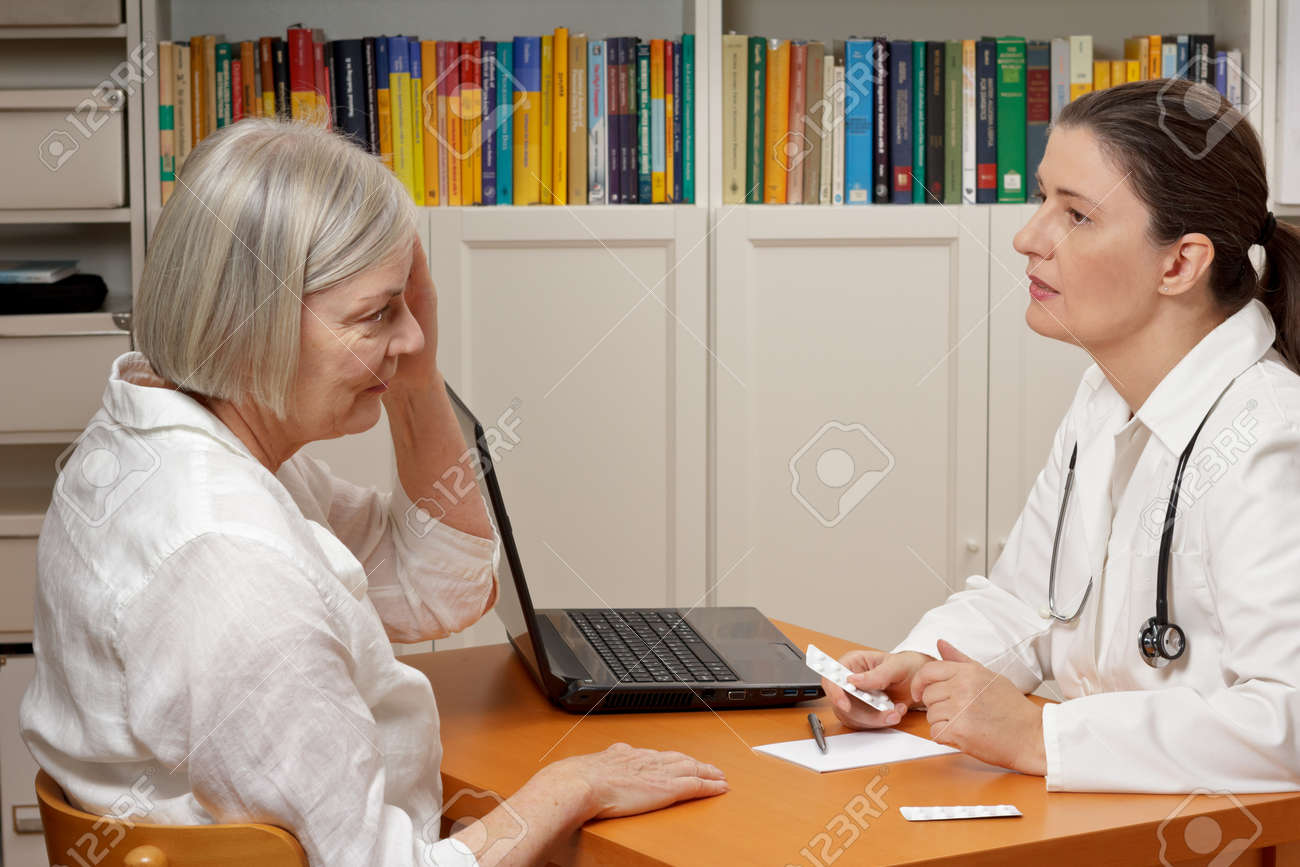 Female middle aged doctor in white coat listening attentively to an old woman with headache or migraine - 65805526 Female middle aged doctor in white coat listening attentively to an old woman with headache or migraine - 65805526