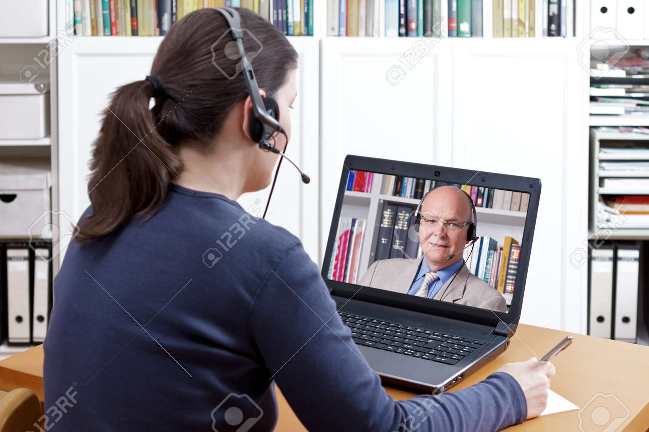 Woman with pen and paper and a headset in front of her laptop making a video call with her friendly professor, text space - 79662354 Woman with pen and paper and a headset in front of her laptop making a video call with her friendly professor, text space - 79662354