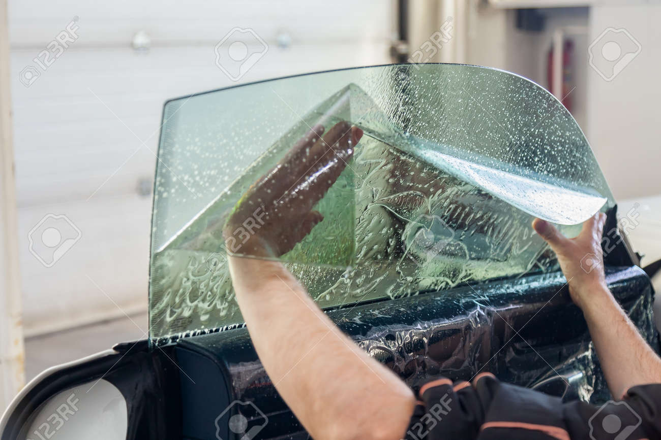 The wizard for installing additional equipment sticks a tint film on the side front glass of the car and flattens it by hand to fit the glass with a greenish tint in the auto service. - 119144410 The wizard for installing additional equipment sticks a tint film on the side front glass of the car and flattens it by hand to fit the glass with a greenish tint in the auto service. - 119144410