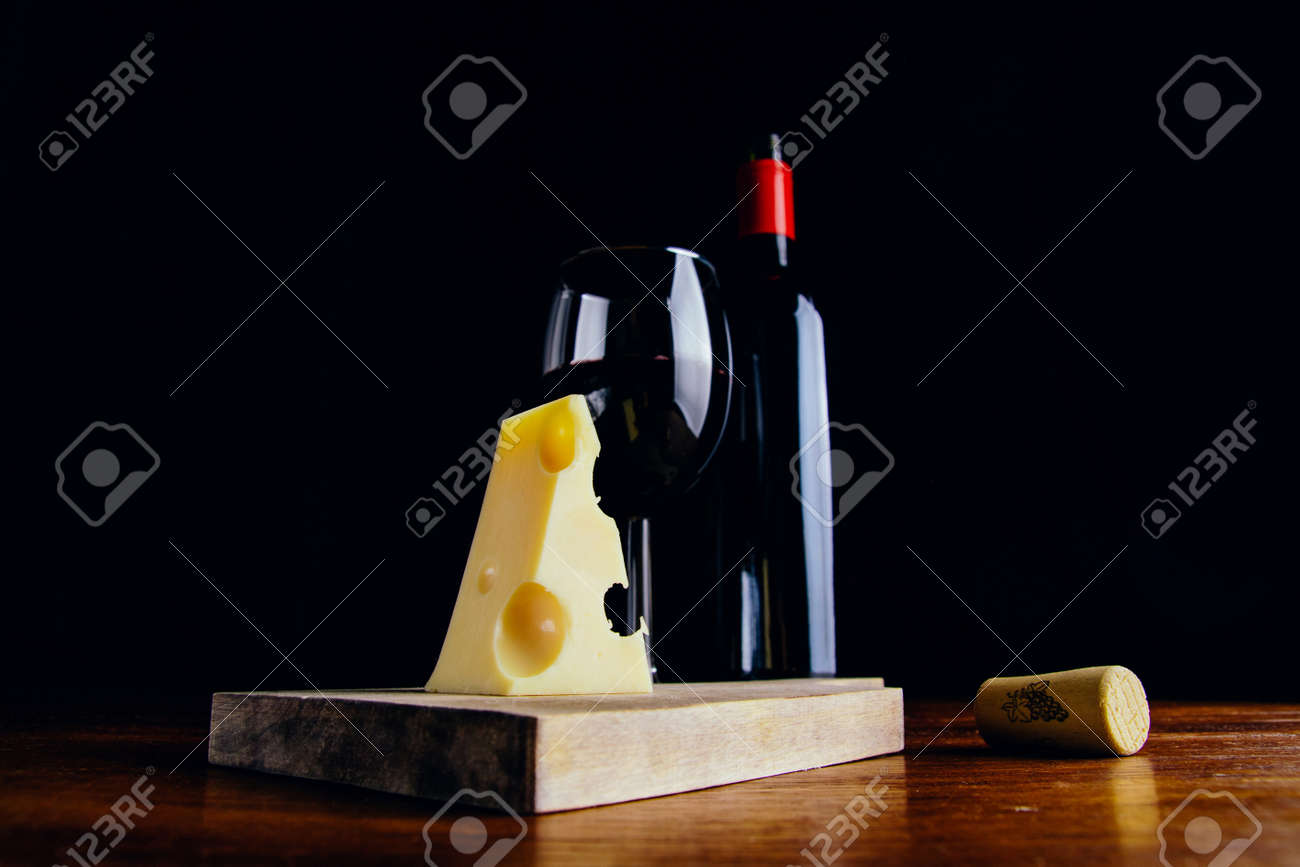 A glass of red wine, a bottle and a piece of cheese on a wooden table and a dark background - 151495021 A glass of red wine, a bottle and a piece of cheese on a wooden table and a dark background - 151495021