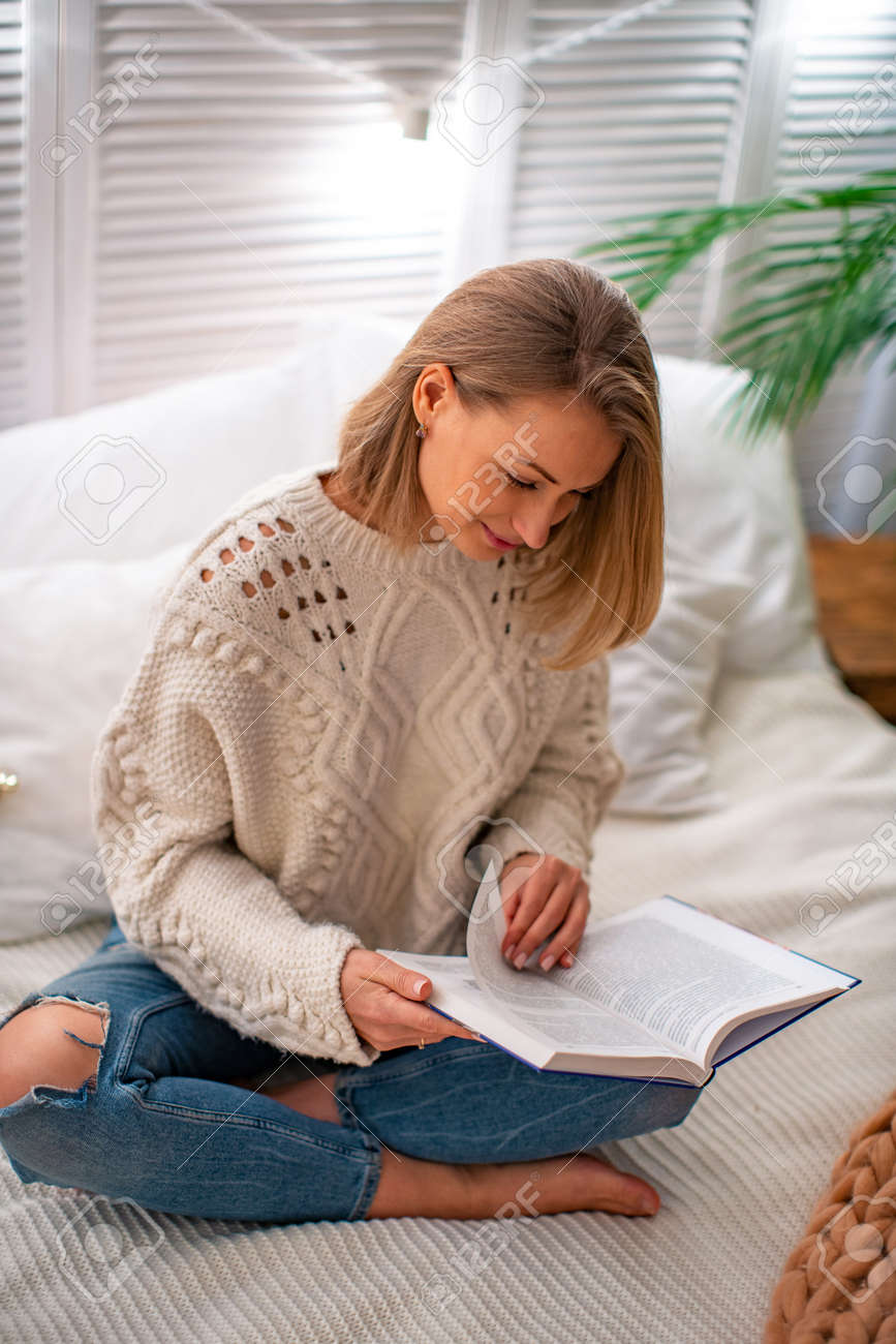 young smiling woman is sitting on the bed with a book. The blonde spends time studying educational literature. The evening reading. The concept of leisure. Stay at home - 145717931 young smiling woman is sitting on the bed with a book. The blonde spends time studying educational literature. The evening reading. The concept of leisure. Stay at home - 145717931