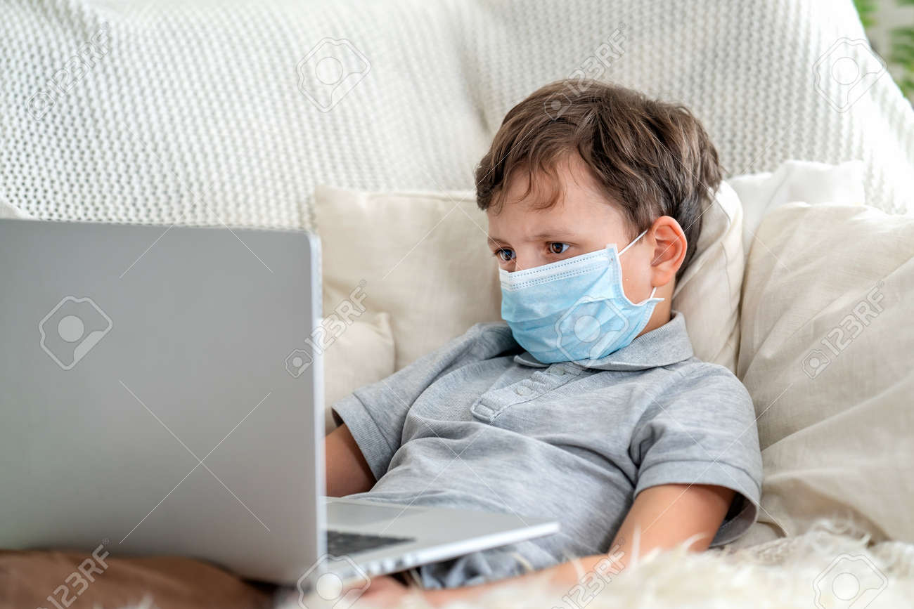 Focused boy in a mask, lying on the couch with a laptop during the quarantine period. He does not go to school, is in isolation, on distance learning. Stay at home, e-learning. - 145987788 Focused boy in a mask, lying on the couch with a laptop during the quarantine period. He does not go to school, is in isolation, on distance learning. Stay at home, e-learning. - 145987788