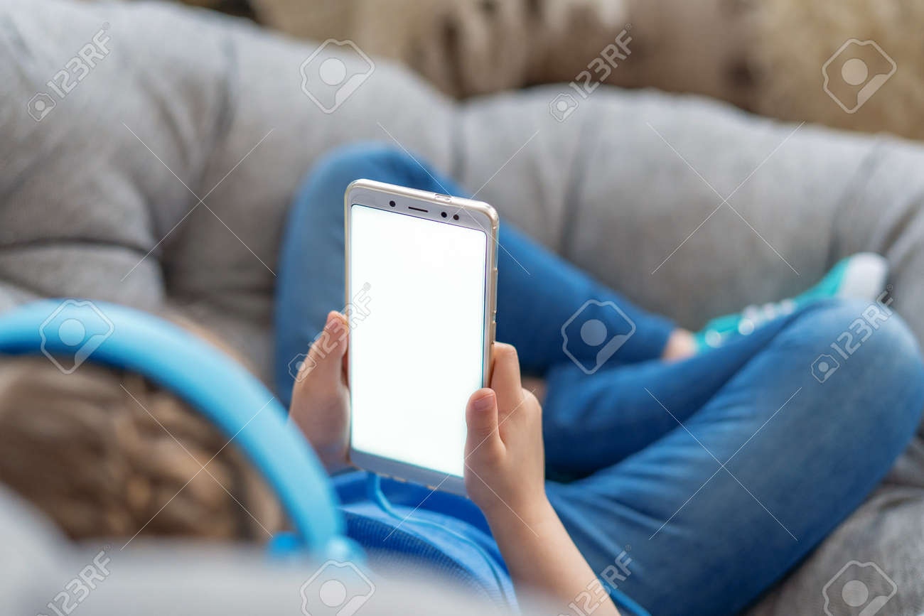 little girl with headphones and holding smartphone white screen in her hands is lying on sofa and looking at mobile phone screen. Close up. Selective focus on hand. Concept leisure during quarantine - 146984507 little girl with headphones and holding smartphone white screen in her hands is lying on sofa and looking at mobile phone screen. Close up. Selective focus on hand. Concept leisure during quarantine - 146984507