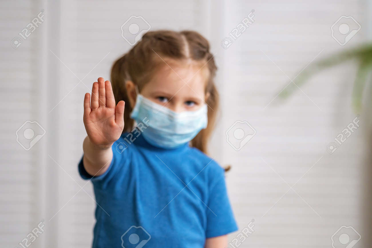 A little girl in a medical mask looks at the camera and makes a stop gesture during the coronavirus pandemic on a light background. The child puts his hand forward to determine the social distance. - 146984510 A little girl in a medical mask looks at the camera and makes a stop gesture during the coronavirus pandemic on a light background. The child puts his hand forward to determine the social distance. - 146984510