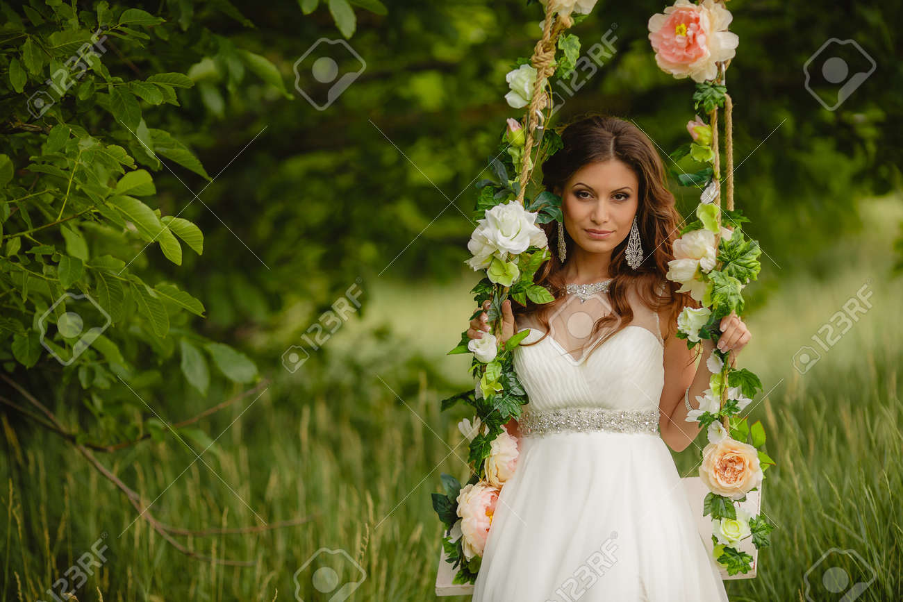 スイングでスイング白いウェディング ドレスの若い美しい花嫁薔薇をあしらった夏の緑豊かな公園 ピアス 左手ブレスレット 茶色の目 長い巻き毛の上のあなたの耳であなたの結婚指輪を着用します の写真素材 画像素材 Image