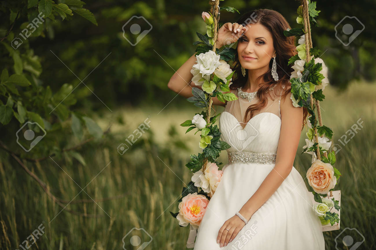 ブランコに揺れて白いウェディング ドレスで若い美しい花嫁薔薇をあしらった夏に緑豊かな公園 左手ブレスレット 茶色の目と長い巻き毛の髪に 耳にピアスであなたの結婚指輪を身に着けています の写真素材 画像素材 Image
