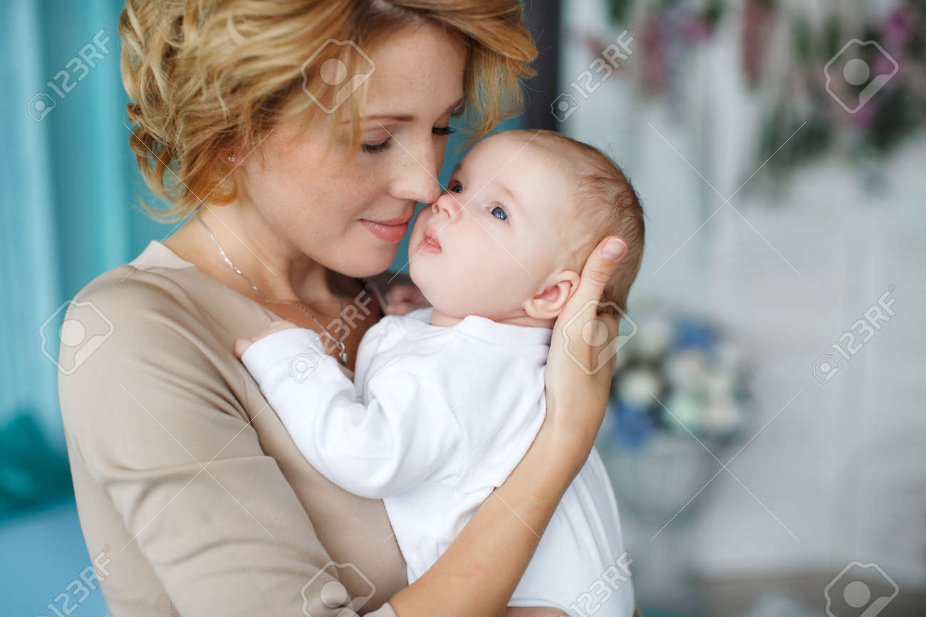 Jeune Femme Belle Avec Des Cheveux Blonds Epais Vetue D Une Robe De Couleur Beige Porte Autour De Son Cou Une Chaine En Argent Avec Un Pendentif Tenant Son Fils Nouveau Ne Le Bebe