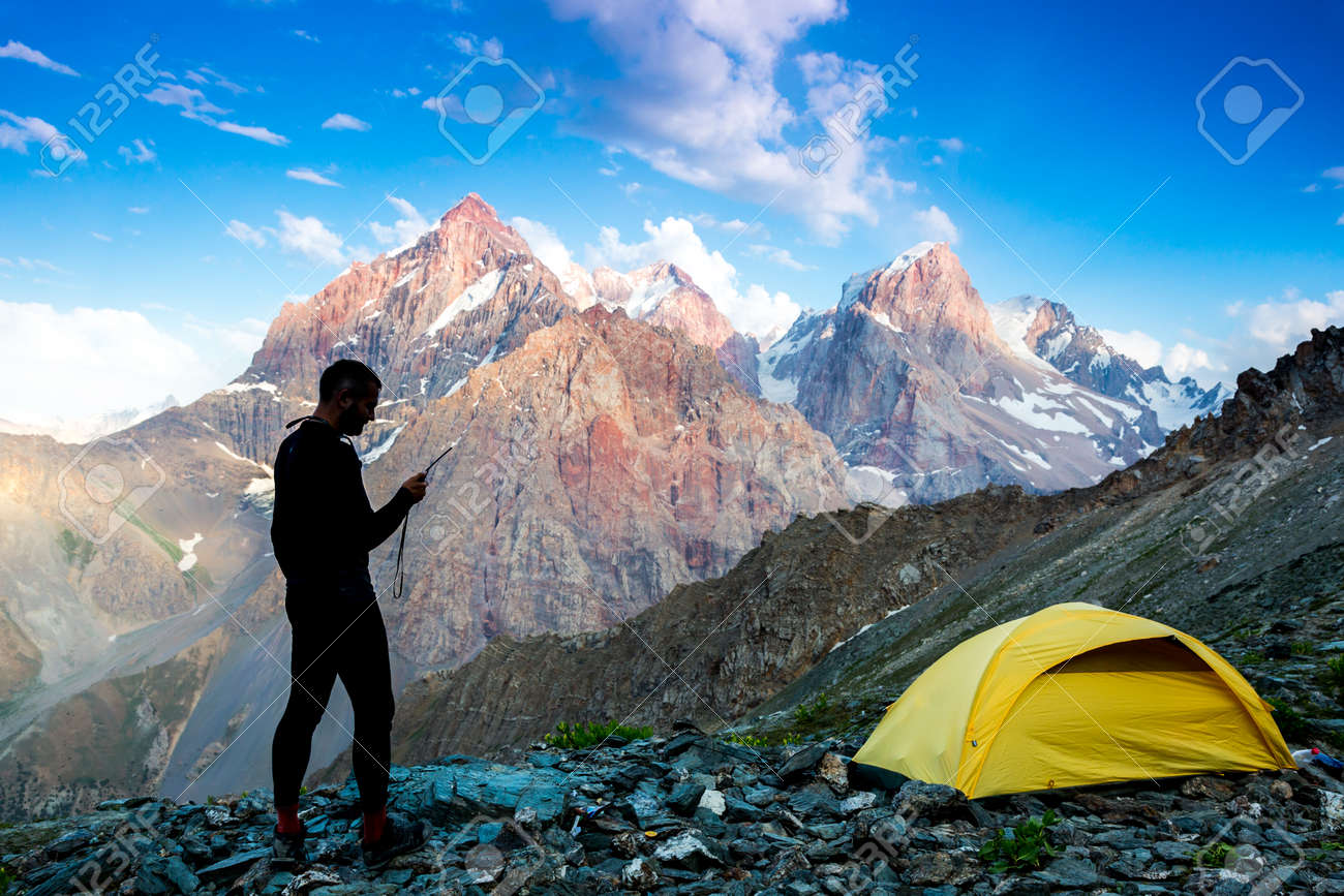 Silhouette of man in wild mountain landscape walk along yellow camping tent holding radio transmitter connection with team blue sky sunny evening - 51765590 Silhouette of man in wild mountain landscape walk along yellow camping tent holding radio transmitter connection with team blue sky sunny evening - 51765590