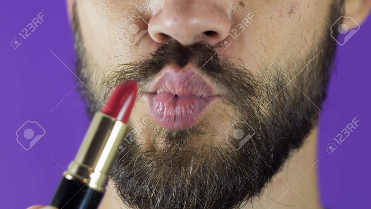 A young guy with a beard paints his lips with red lipstick. Close-up of a bearded man, he painted lips with bright lipstick. A bearded man puts red lipstick on his lips, smiles and smacks his lips. - 152947780 A young guy with a beard paints his lips with red lipstick. Close-up of a bearded man, he painted lips with bright lipstick. A bearded man puts red lipstick on his lips, smiles and smacks his lips. - 152947780