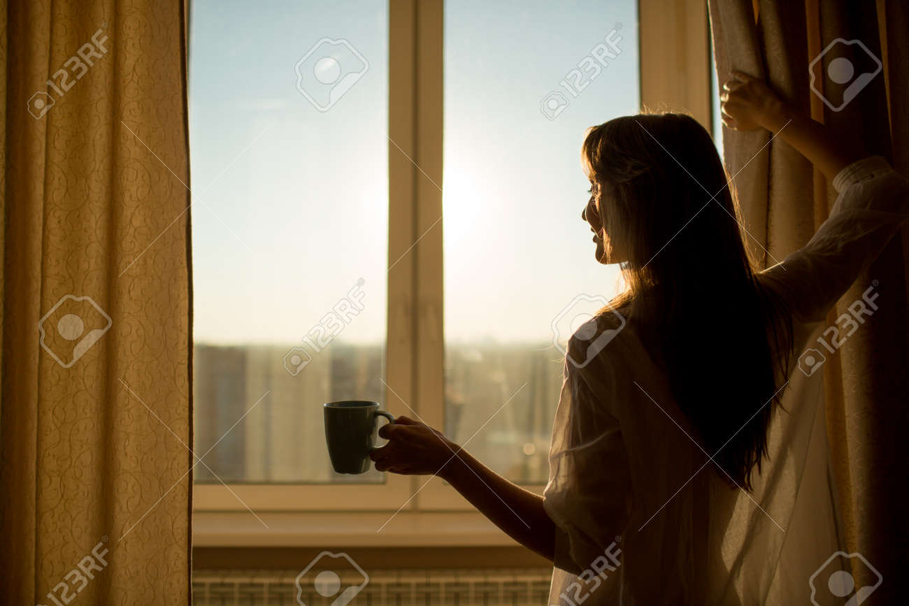 Woman in the morning. Attractive woman with neat body is holding a cup with hot tea or coffee and looking at the sunrise standing near the window in her home and having a perfect cozy morning. - 31878159 Woman in the morning. Attractive woman with neat body is holding a cup with hot tea or coffee and looking at the sunrise standing near the window in her home and having a perfect cozy morning. - 31878159