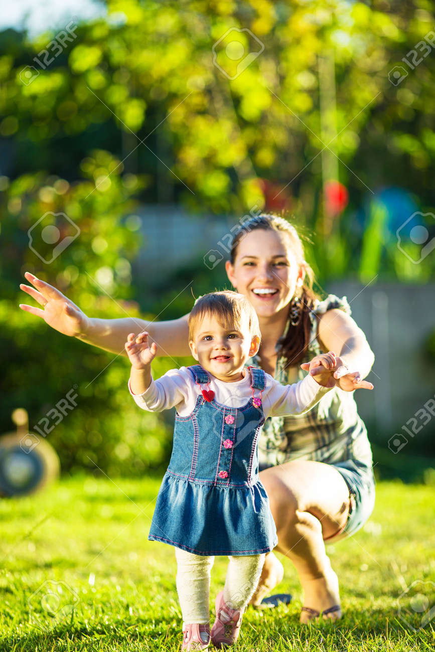 Baby girl is doing her first steps with mothers help. Cute little girl learns to walk with her young mom helping her in the sunny garden outdoors. Happy childhood and motherhood concept. - 32330490 Baby girl is doing her first steps with mothers help. Cute little girl learns to walk with her young mom helping her in the sunny garden outdoors. Happy childhood and motherhood concept. - 32330490