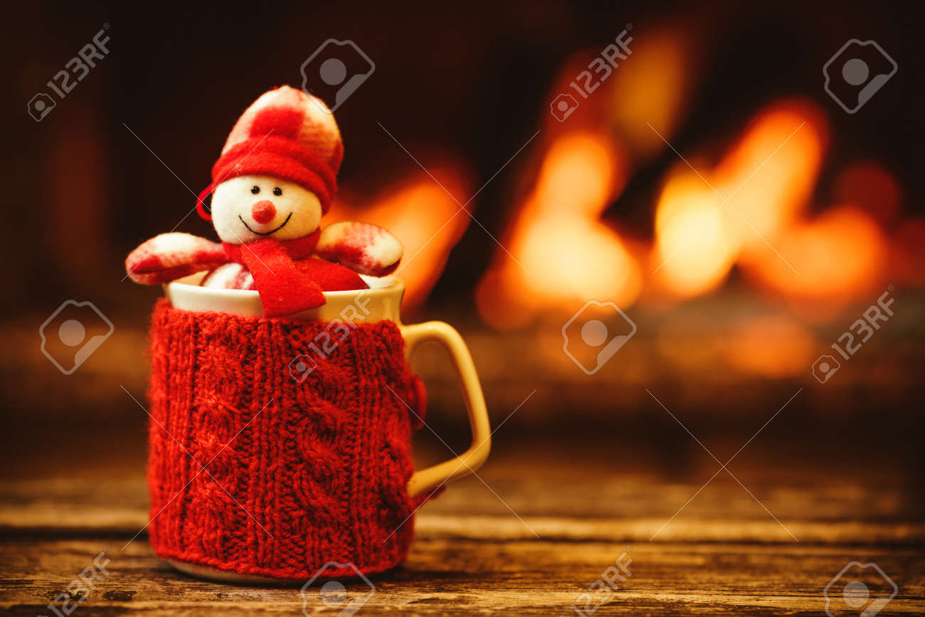 Cup of hot drink in front of warm fireplace. Holiday Christmas concept. Mug in red knitted mitten, decorated with snowman toy, standing near fireside. Cozy relaxed magical atmosphere in a chalet. - 46927099 Cup of hot drink in front of warm fireplace. Holiday Christmas concept. Mug in red knitted mitten, decorated with snowman toy, standing near fireside. Cozy relaxed magical atmosphere in a chalet. - 46927099