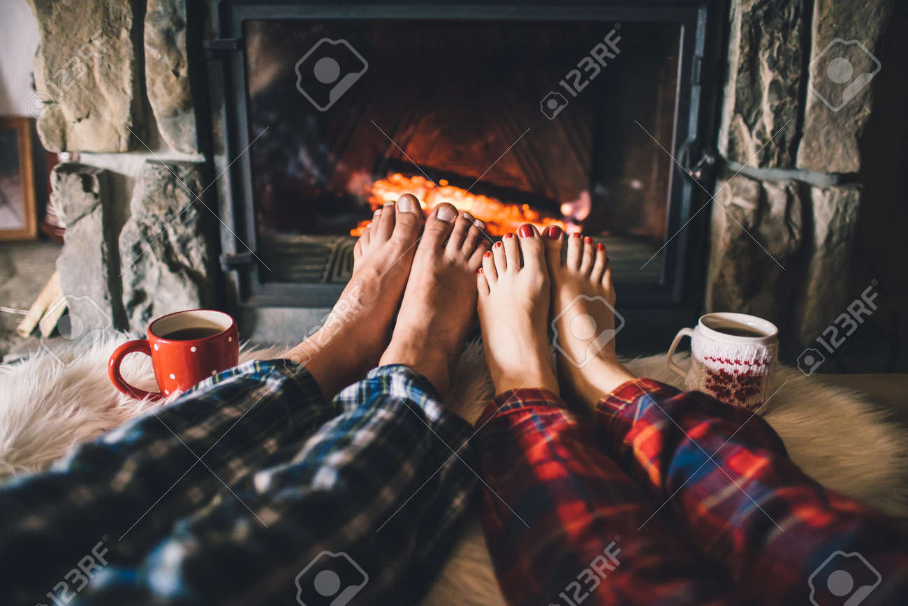Man and Woman relaxes by warm fire with a cup of hot drink and warming up her feet. - 58947692 Man and Woman relaxes by warm fire with a cup of hot drink and warming up her feet. - 58947692