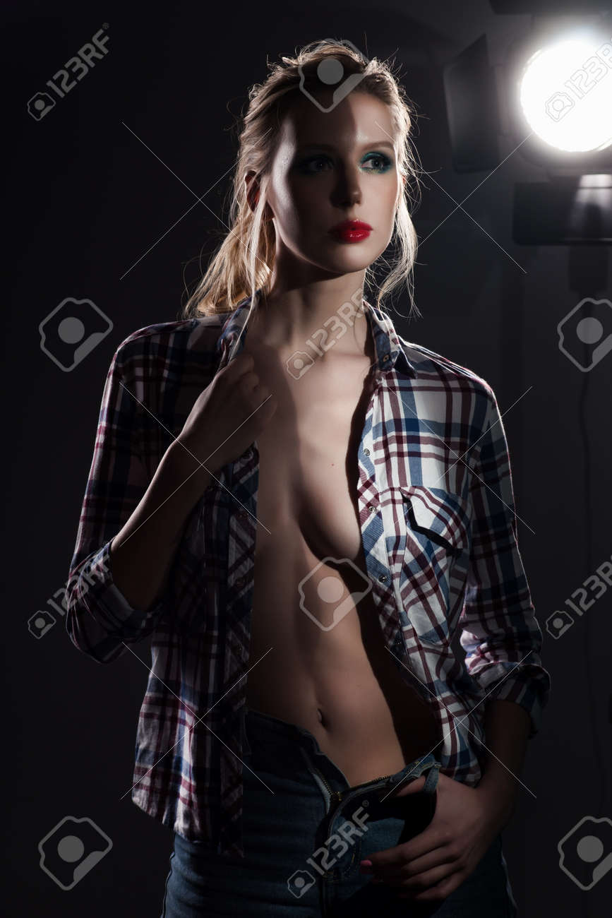 slender big tits A Beautiful Slender Blonde Big Breasts Girl Without A Bra Wearing An Unbuttoned Checkered Shirt And Jeans Half Covered With Vertical Shadow Near The Studio Lamp. Copy Space. Grey Background. Stock Photo,