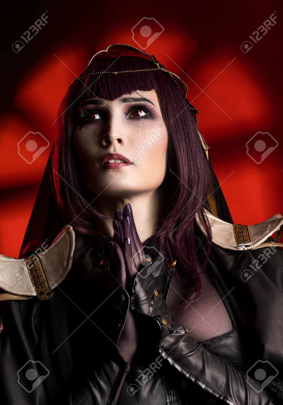 A Beautiful Busty Cosplay Girl Wearing An Erotic Leather Costume Sensually  Cries With Tears In Her Face And Makes A Praying Gesture On A Dark And Red  Background. Close Up Portrait Stock