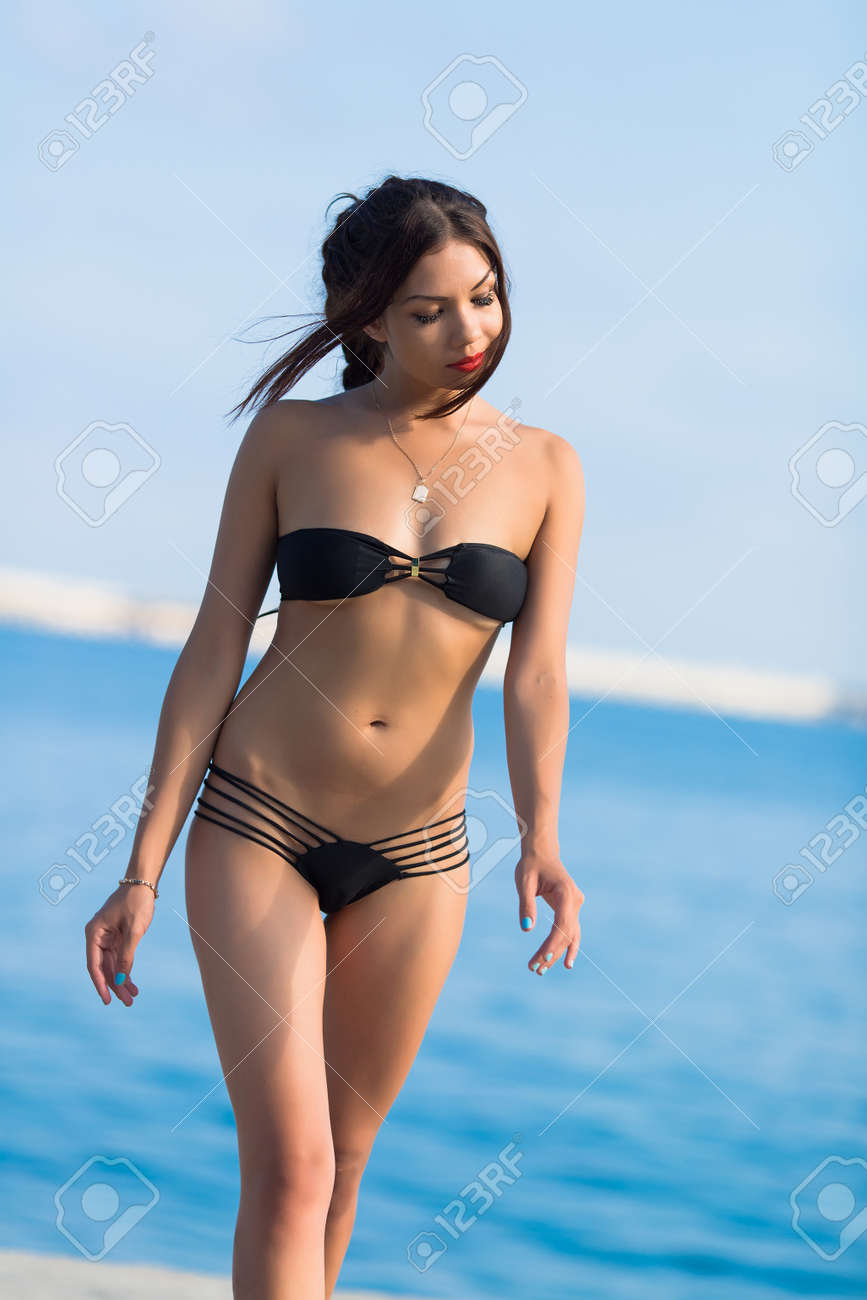 下へ見ている海岸に沿って歩く黒いビキニで魅力的な若い女性 の写真素材 画像素材 Image