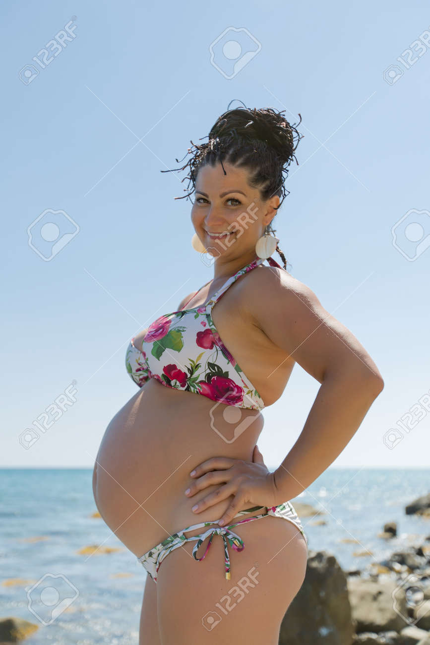 海に対して提起するビキニの妊娠中の女性 笑みを浮かべてカメラを見て丁拳銃水着立っている腕の中で魅力的な妊婦 の写真素材 画像素材 Image