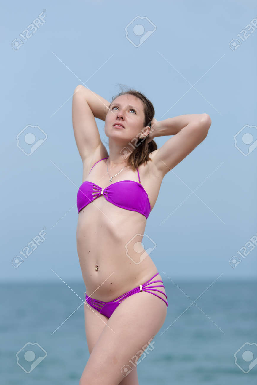 見上げる水着モデル 海に対して腕を上げて立っている紫色のビキニの若い女性の肖像画。曇りの日に見上げるビーチでポーズをとって別の水着の女の子の写真素材・画像素材 Image  172088240