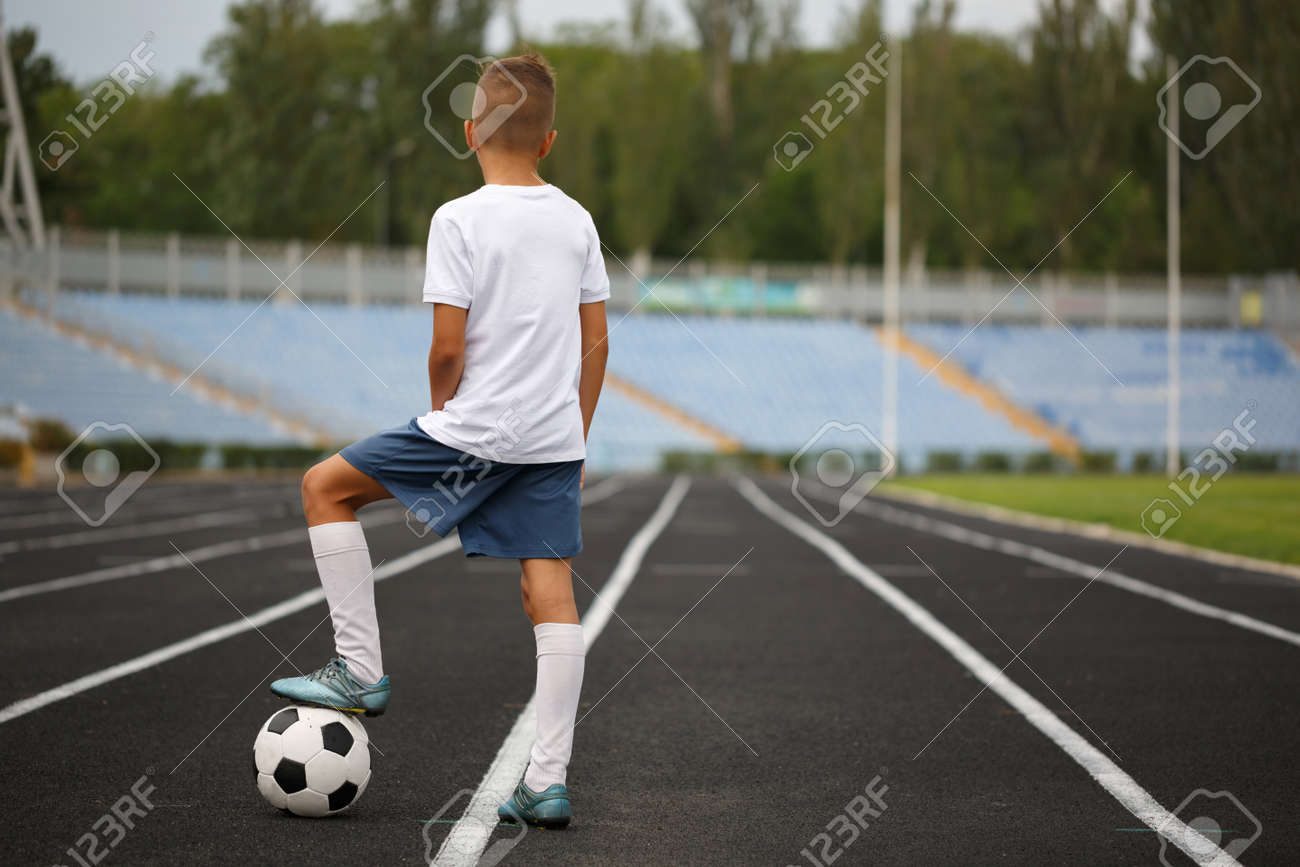 ショート パンツやトレーナー スタジアムでのサッカー ボールの上に 足で愛らしいサッカー プレーヤーのフルレングスの写真 自然の背景にアスファルトで舗装されたパスでボールを持つ少年 の写真素材 画像素材 Image