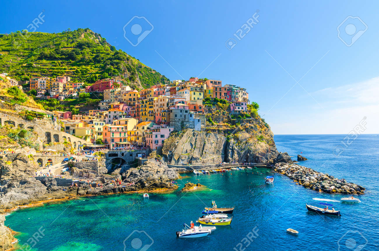 Manarola traditional typical Italian village in National park Cinque Terre, colorful multicolored buildings houses on rock cliff, fishing boats on water, blue sky background, La Spezia, Liguria, Italy - 123537178 Manarola traditional typical Italian village in National park Cinque Terre, colorful multicolored buildings houses on rock cliff, fishing boats on water, blue sky background, La Spezia, Liguria, Italy - 123537178