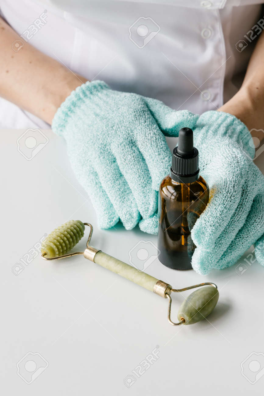 Brown plastic bottle for liquid cosmetics in womans hands in green gloves, green jade massage roller on white table. Cosmetologist holding spin care products. - 152134220 Brown plastic bottle for liquid cosmetics in womans hands in green gloves, green jade massage roller on white table. Cosmetologist holding spin care products. - 152134220