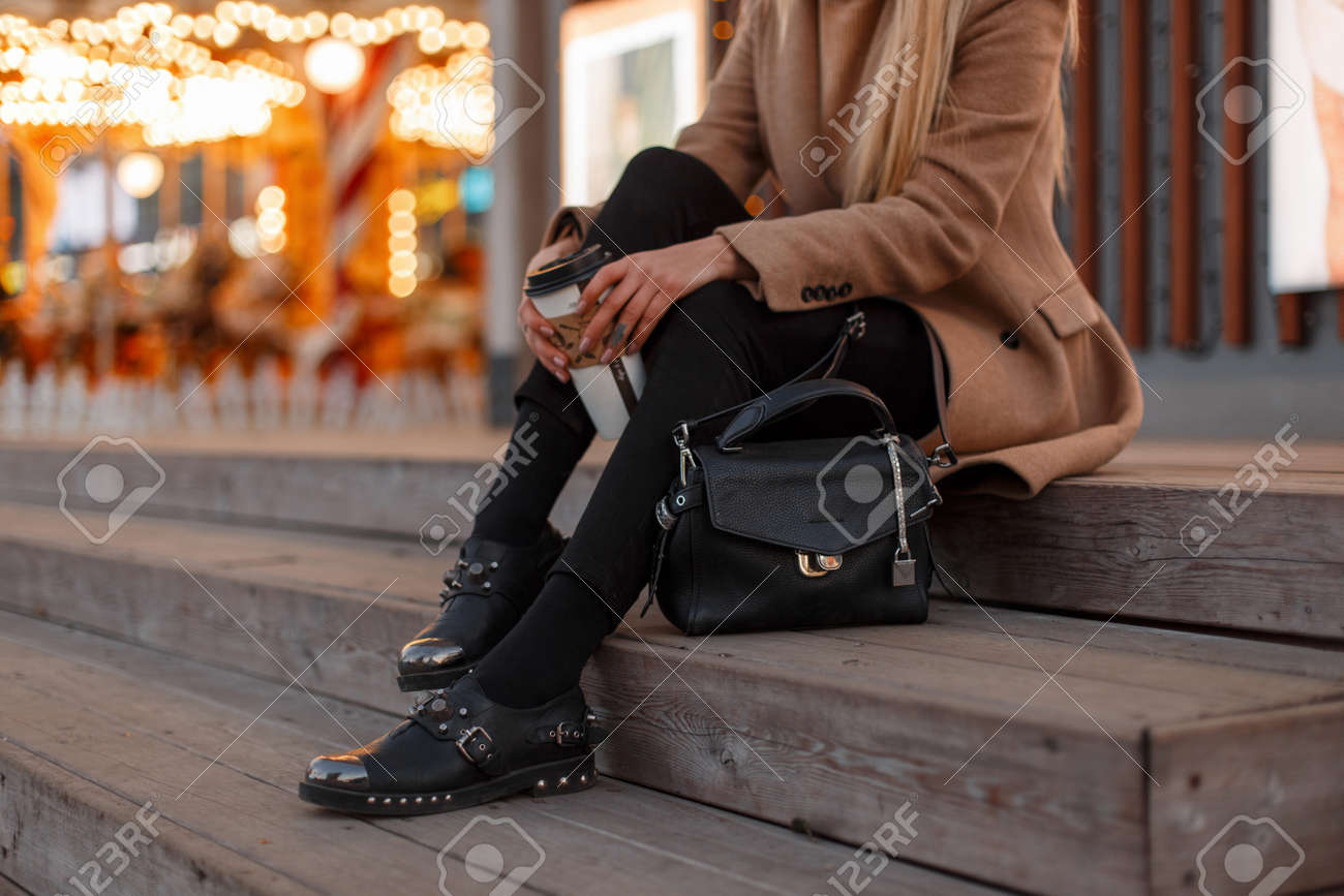 Young girl in a vintage coat with a warm knitted sweater, stylish jeans, black leather fashionable modern shoes and a stylish leather bag sits with a cup of coffee. Women's stylish clothing. Close-up - 112760418 Young girl in a vintage coat with a warm knitted sweater, stylish jeans, black leather fashionable modern shoes and a stylish leather bag sits with a cup of coffee. Women's stylish clothing. Close-up - 112760418