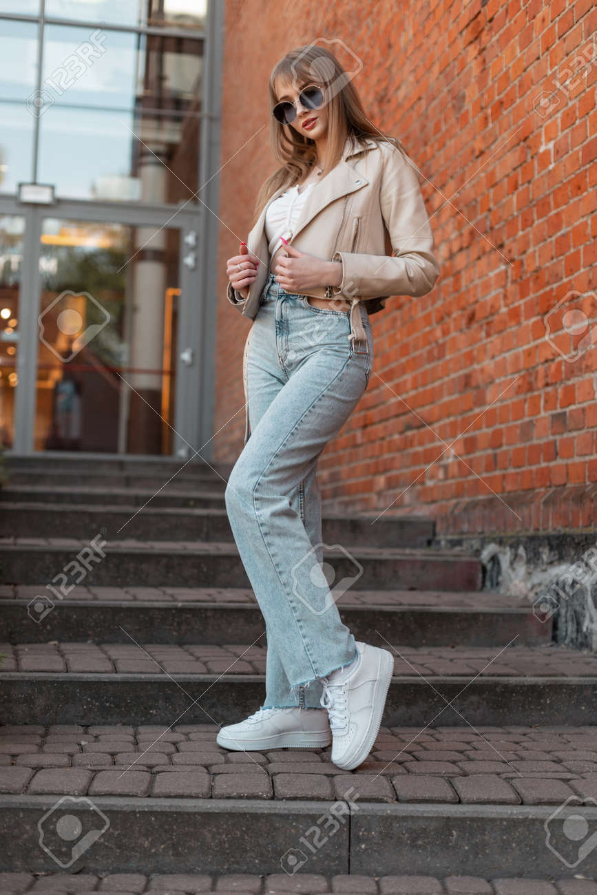 Hermosa Joven Modelo Hipster Con Gafas De Sol De Moda En Ropa Casual De Moda Con Chaqueta De Cuero De Moda, Jeans De Cintura Alta Y Zapatillas Caminando Cerca De Un