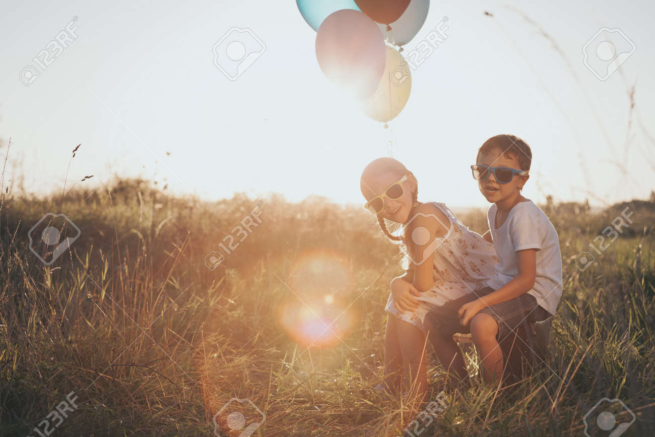 Criancas Pequenas Felizes Que Jogam Na Estrada No Tempo Do Por Do Sol Eles Sentados Na Mala No Parque E Segurando Baloes Na Mao Criancas Se Divertindo Na Natureza Conceito De Felicidade