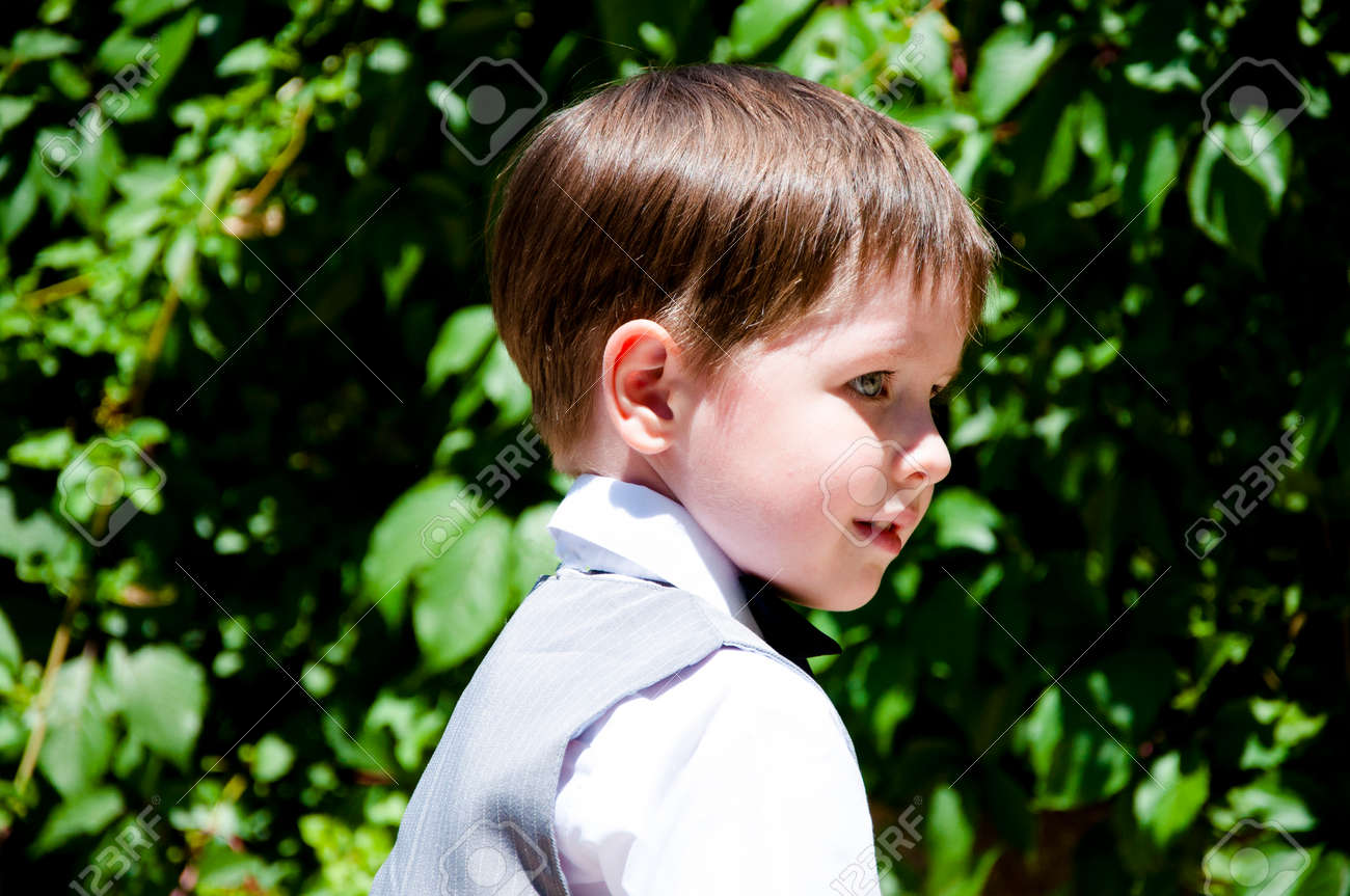 Bonito Pequeno Menino Ou Cavalheiro Crianca Com Sorrindo Em Formal Terno Cinza De Colete Camisa Branca E Gravata Preta Ensolarada Ao Ar Livre No Fundo Da Parede Pedregoso Com Plantas De Hera