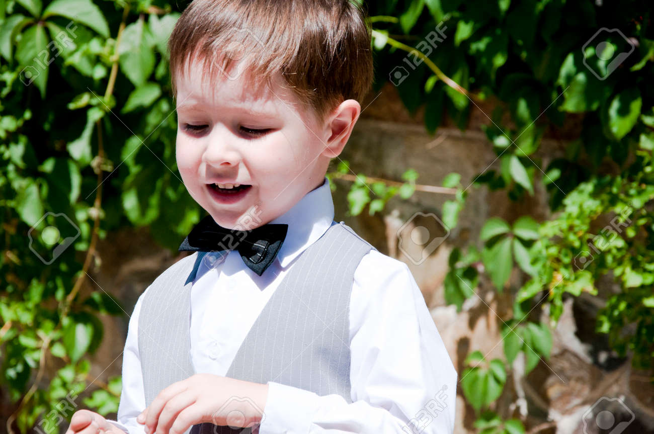 Bonito Pequeno Menino Ou Cavalheiro Crianca Com Sorrindo Em Formal Terno Cinza De Colete Camisa Branca E Gravata Preta Ensolarada Ao Ar Livre No Fundo Da Parede Pedregoso Com Plantas De Hera