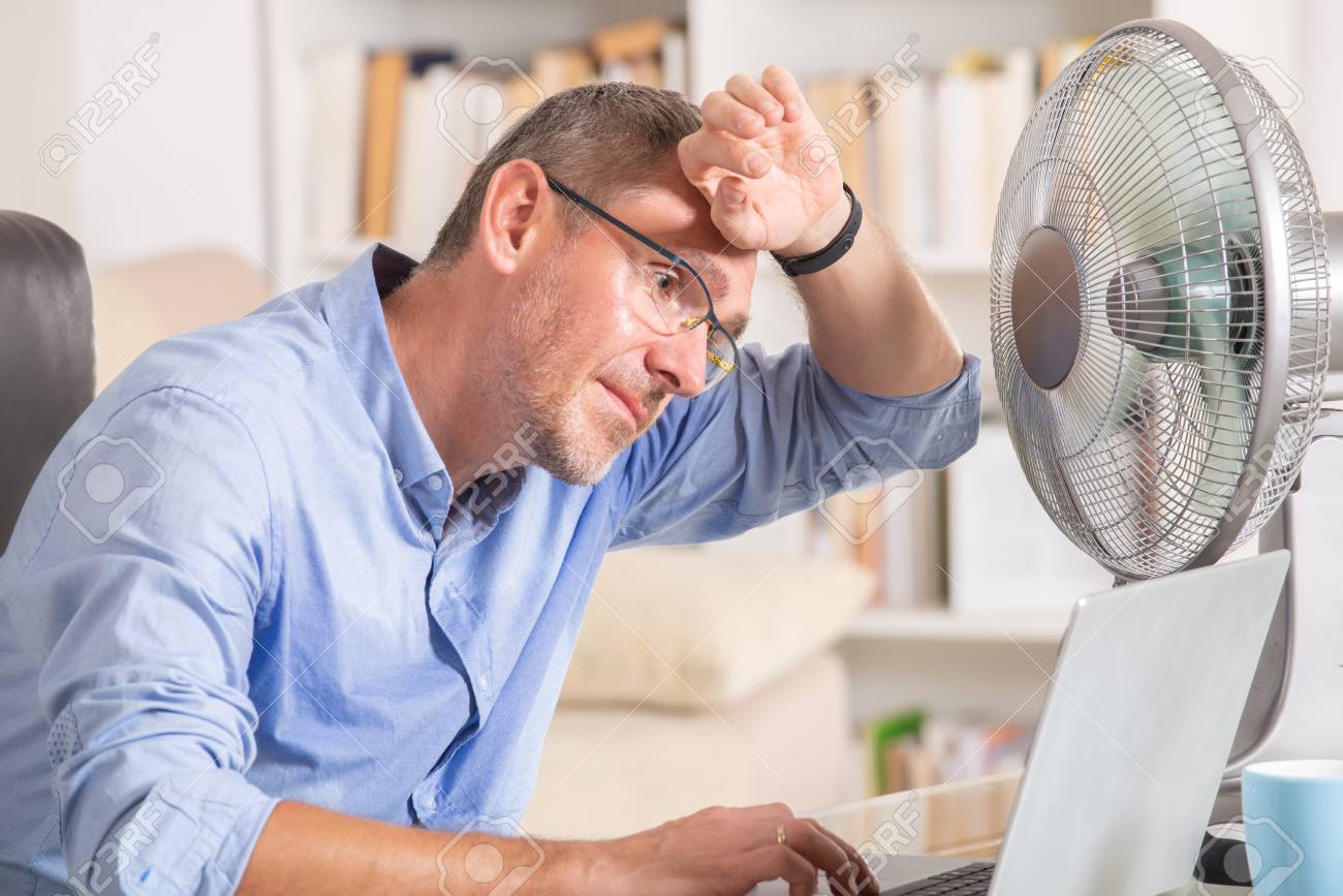 Man suffers from heat while working in the office and tries to cool off by the fan - 104782940 Man suffers from heat while working in the office and tries to cool off by the fan - 104782940