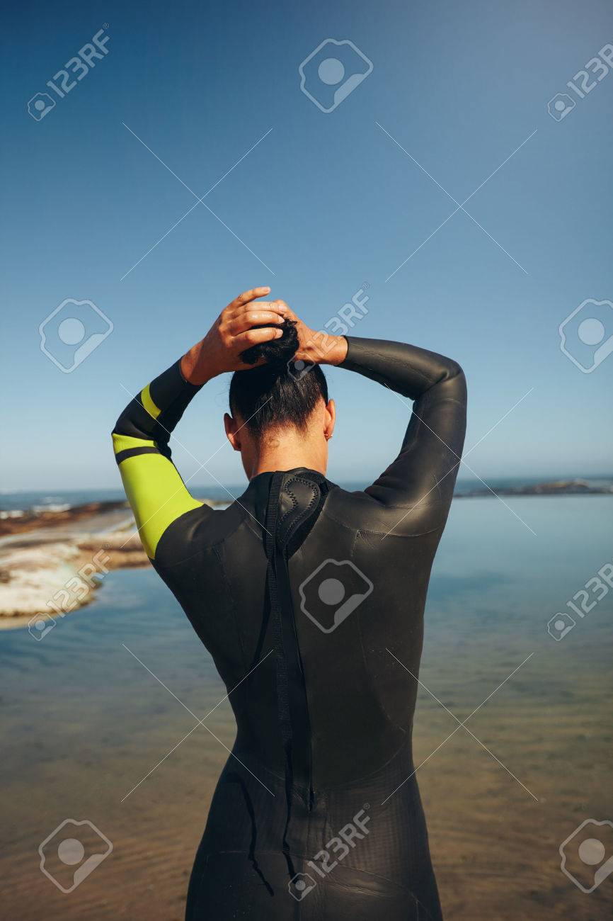 ウェット スーツを着てレースに準備して湖の上の若いトライアスリートの背面 女子トライアスロン選手彼女の髪を結び 競争のための準備します の写真素材 画像素材 Image