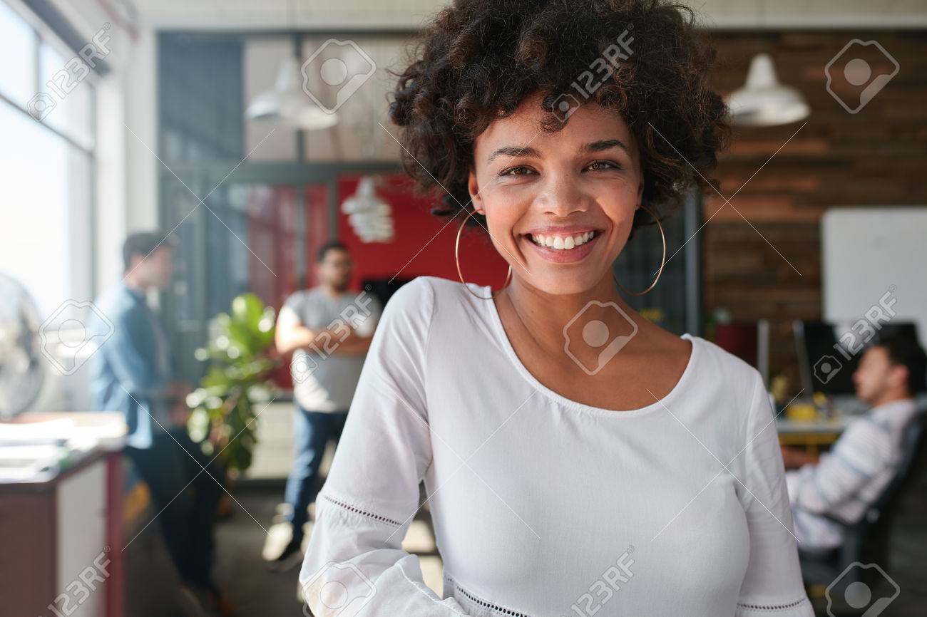 Portrait of smiling young african businesswoman with people in background. Cheerful young woman standing relaxed in her office, looking at camera and smiling. - 51770057 Portrait of smiling young african businesswoman with people in background. Cheerful young woman standing relaxed in her office, looking at camera and smiling. - 51770057