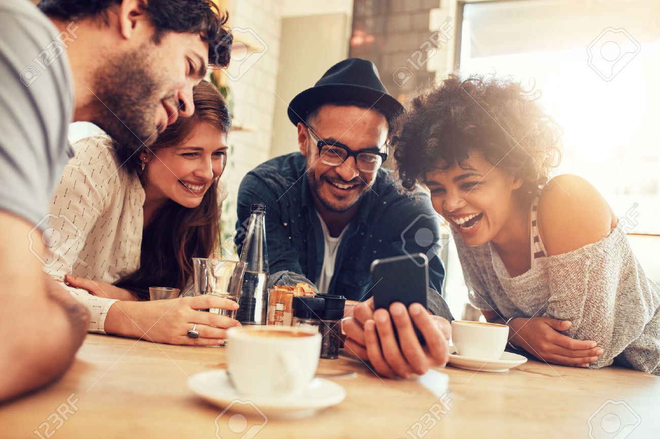 Portrait of cheerful young friends looking at smart phone while sitting in cafe. Mixed race people sitting at a table in restaurant using mobile phone. - 52407809 Portrait of cheerful young friends looking at smart phone while sitting in cafe. Mixed race people sitting at a table in restaurant using mobile phone. - 52407809
