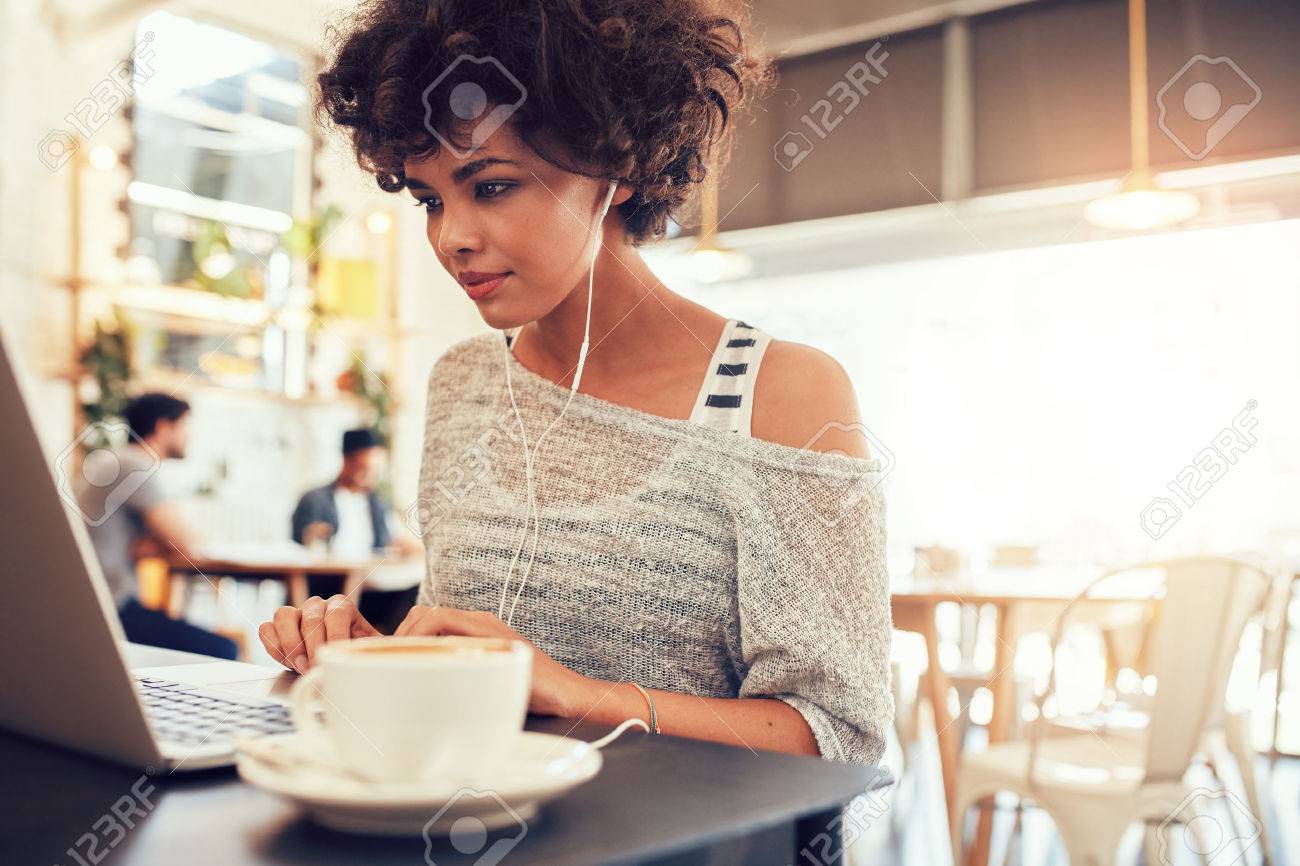 Portrait of an attractive young woman with earphones using laptop at a cafe. African american woman working on laptop computer at a coffee shop. - 53227907 Portrait of an attractive young woman with earphones using laptop at a cafe. African american woman working on laptop computer at a coffee shop. - 53227907