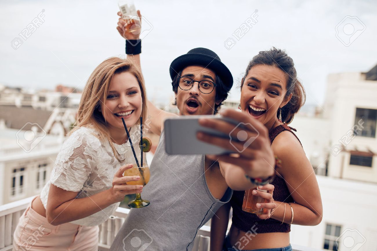 Excited young people taking self portrait with mobile phone during a party. Happy young man and woman taking self portrait at rooftop party. Multiracial people having fun in party with drinks. - 55352771 Excited young people taking self portrait with mobile phone during a party. Happy young man and woman taking self portrait at rooftop party. Multiracial people having fun in party with drinks. - 55352771