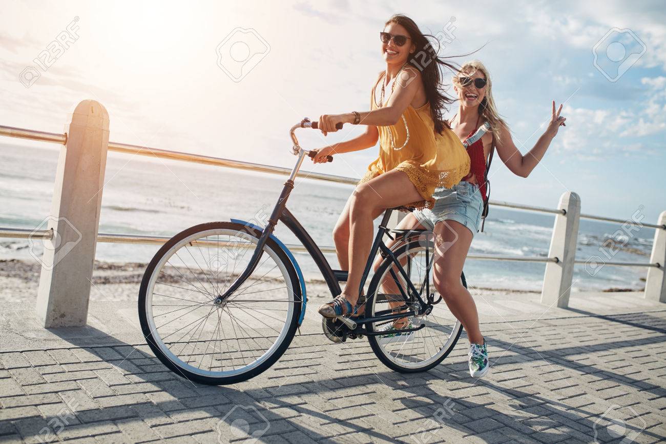 Two stylish young female friends on a bicycle along seaside. Best friends enjoying a day on bike. - 58767243 Two stylish young female friends on a bicycle along seaside. Best friends enjoying a day on bike. - 58767243