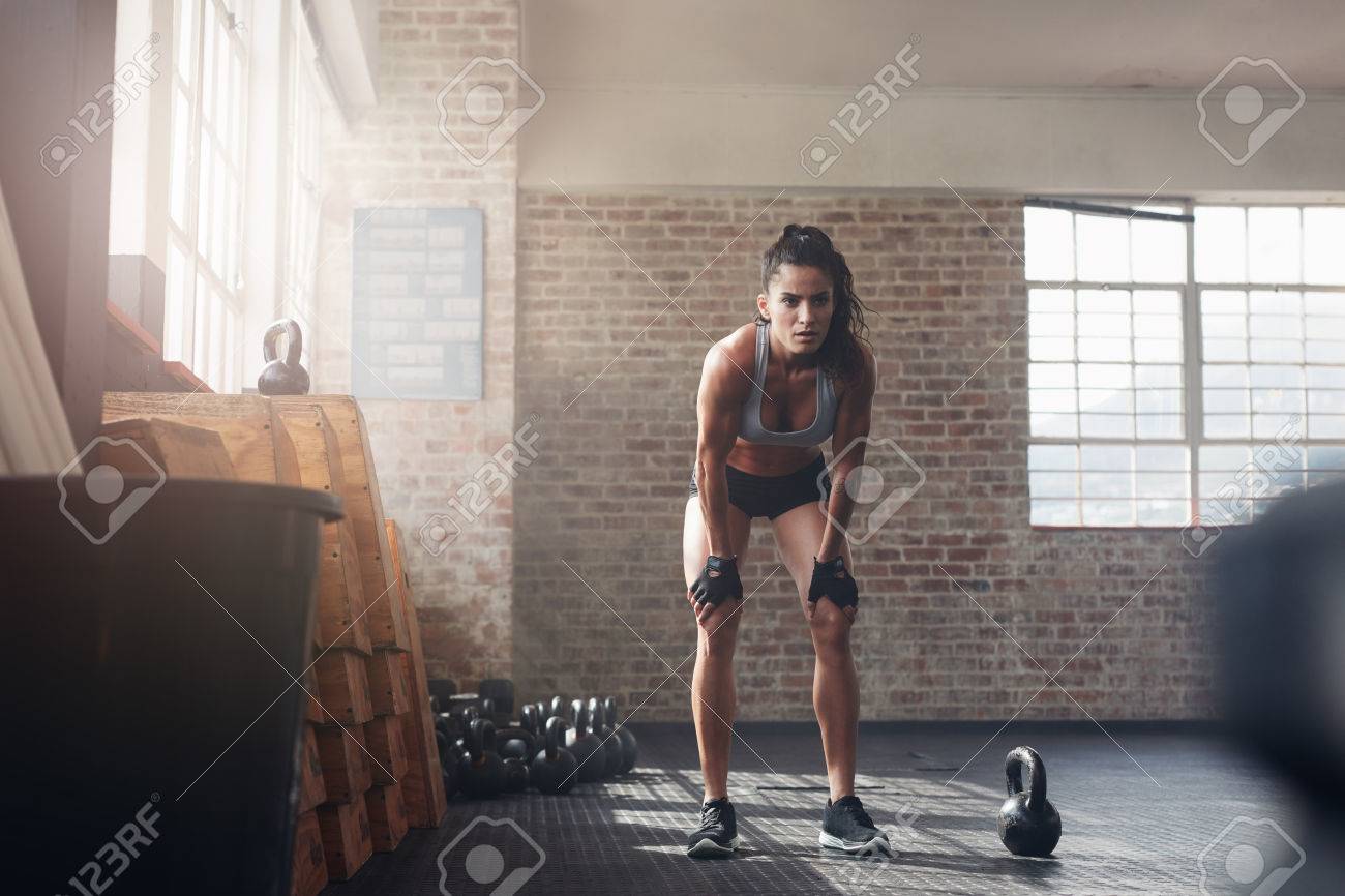 Full length shot of confident young woman at crossfit gym. Muscular sportswoman standing with her hands on knees looking focused about her fitness workout. - 60417857 Full length shot of confident young woman at crossfit gym. Muscular sportswoman standing with her hands on knees looking focused about her fitness workout. - 60417857