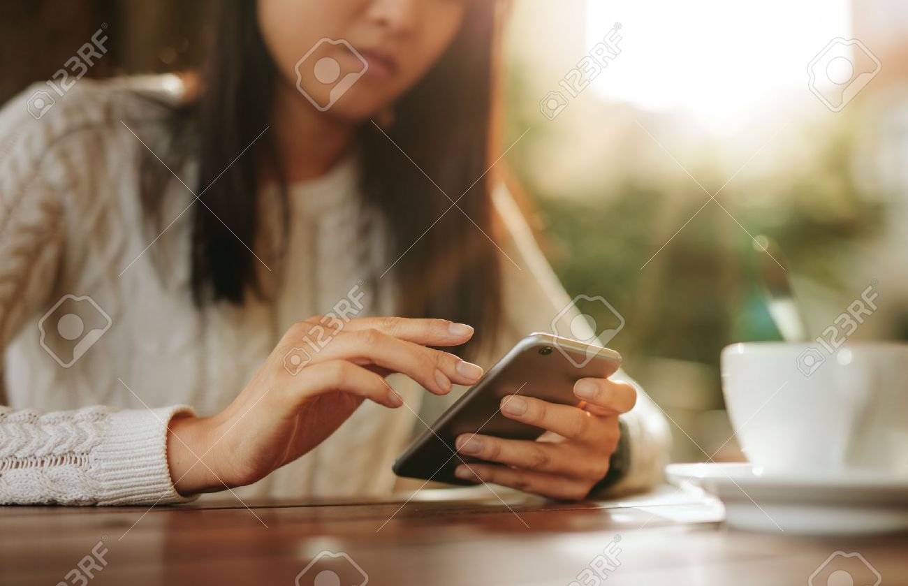 Young asian woman sitting at a table using mobile phone. Smart phone in hands of a female at outdoor cafe. - 62779109 Young asian woman sitting at a table using mobile phone. Smart phone in hands of a female at outdoor cafe. - 62779109