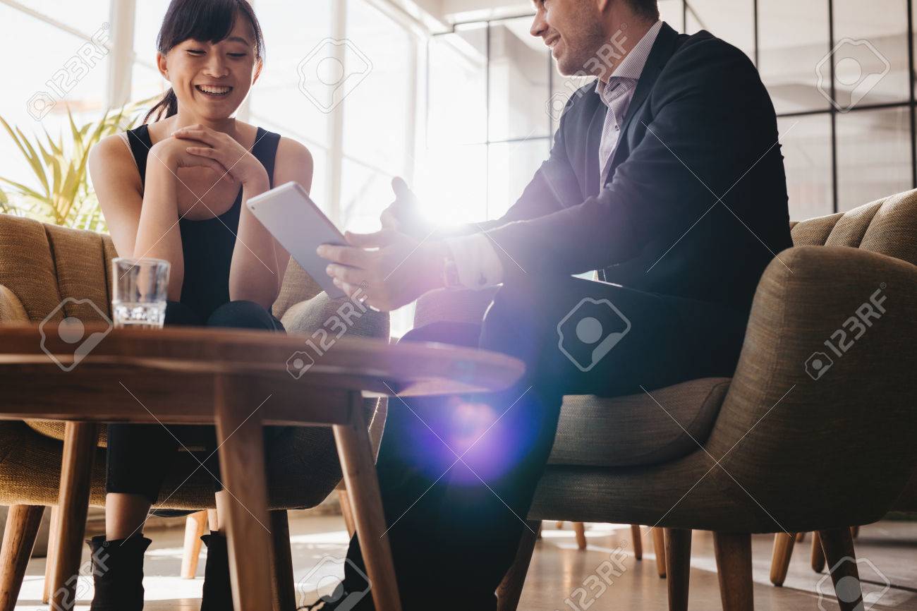 Two corporate colleagues discussing business ideas using digital tablet. Young businessman having meeting with female partner in office lobby - 64920013 Two corporate colleagues discussing business ideas using digital tablet. Young businessman having meeting with female partner in office lobby - 64920013