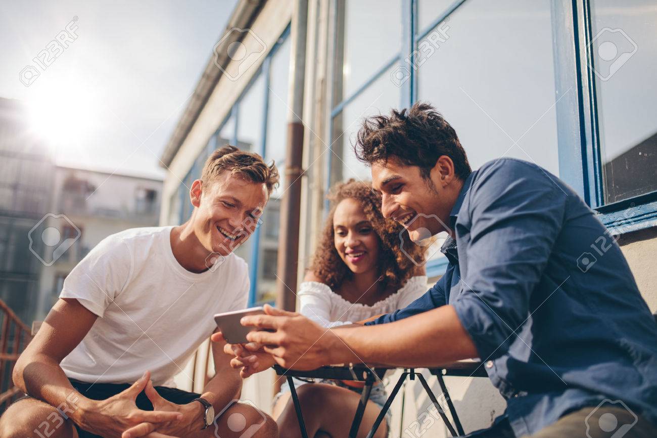 Three young friends sitting outdoors and looking at mobile phone. Group of people sitting at outdoor cafe and watching video on the smartphone. - 64920778 Three young friends sitting outdoors and looking at mobile phone. Group of people sitting at outdoor cafe and watching video on the smartphone. - 64920778