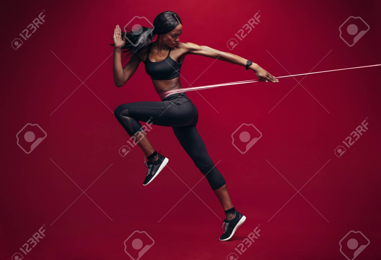 Sporty woman exercising with resistance band on red background. African female athlete working out with elastic bands in studio. - 94183791 Sporty woman exercising with resistance band on red background. African female athlete working out with elastic bands in studio. - 94183791
