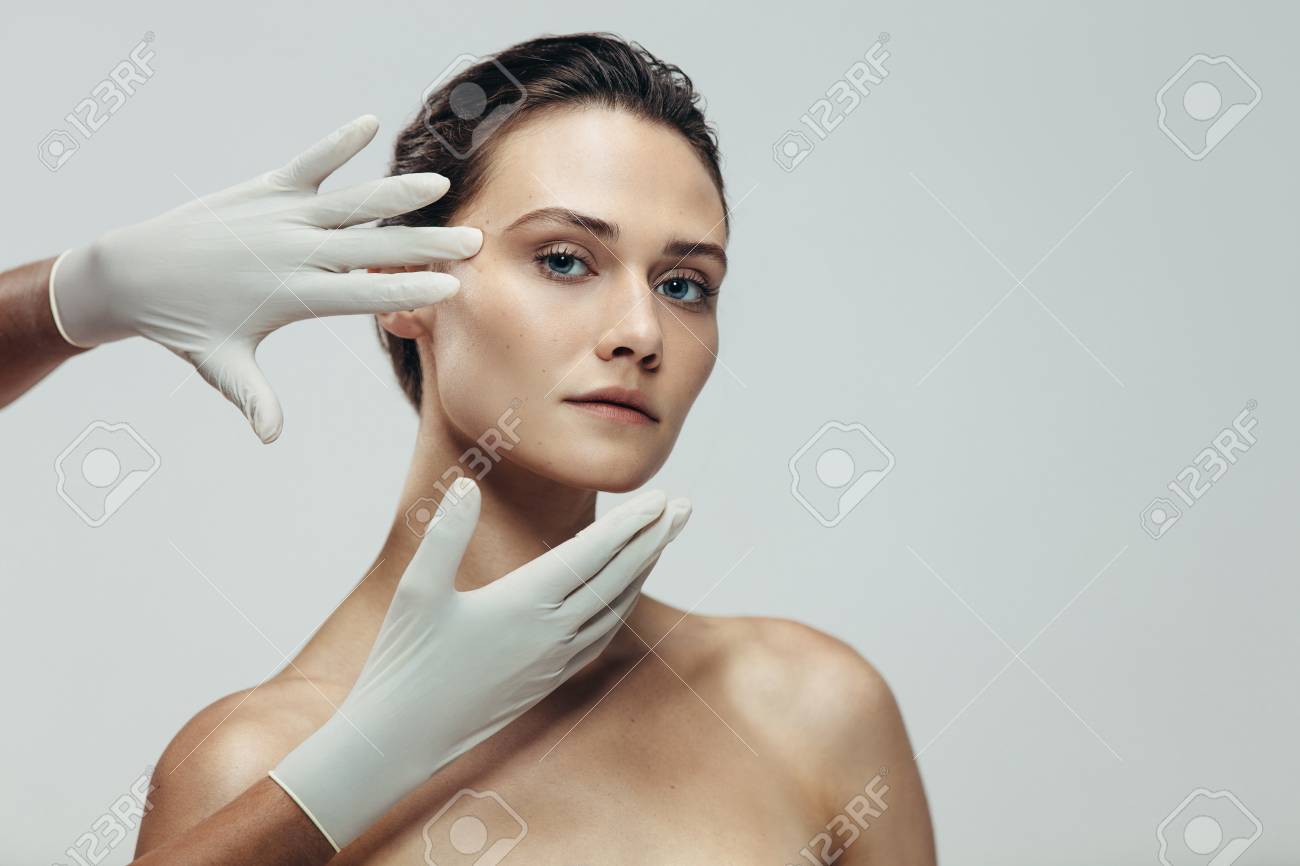 Beautician hands with gloves touching beautiful woman face before plastic surgery. Female standing against grey background with a cosmetologist touching her face. - 109099636 Beautician hands with gloves touching beautiful woman face before plastic surgery. Female standing against grey background with a cosmetologist touching her face. - 109099636