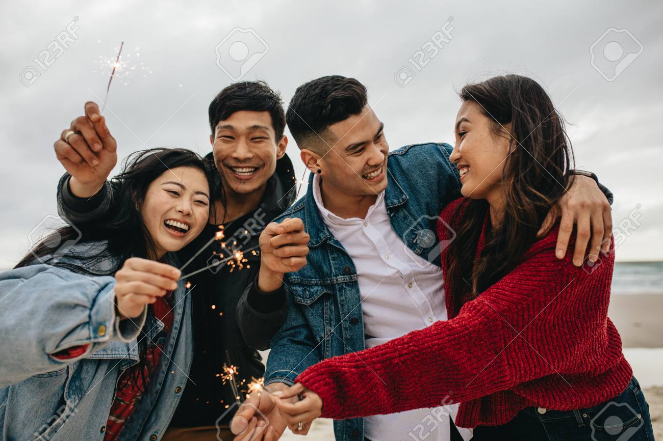 Diverse group of young people celebrating new year's day at the beach. Young asian people having fun with sparklers outdoors at the sea shore. - 119566777 Diverse group of young people celebrating new year's day at the beach. Young asian people having fun with sparklers outdoors at the sea shore. - 119566777